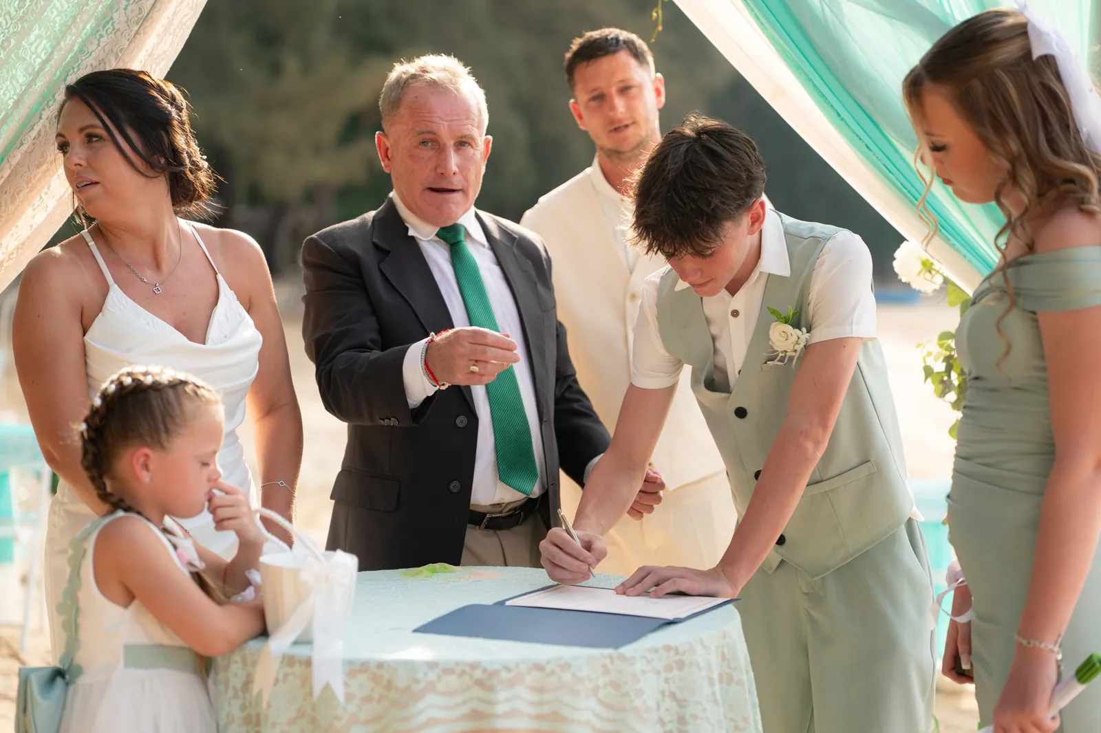 Nicky signing the marriage register at a small table on Hua Beach, Jade beside him and the celebrant looking on with the wedding party around them