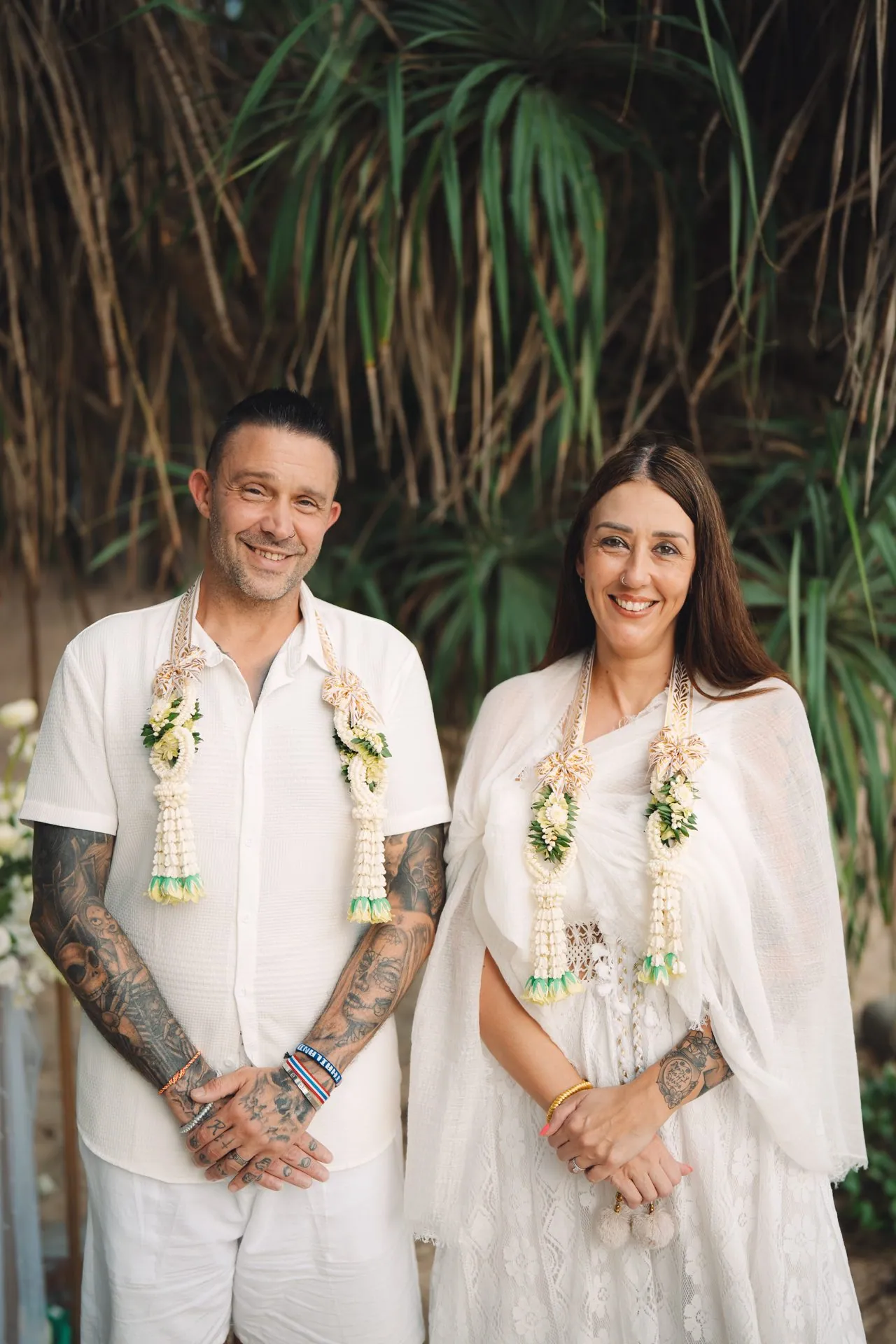 Joanne and Richard standing side by side in white after the ceremony, wearing floral garlands, framed by the trees at Layan Beach