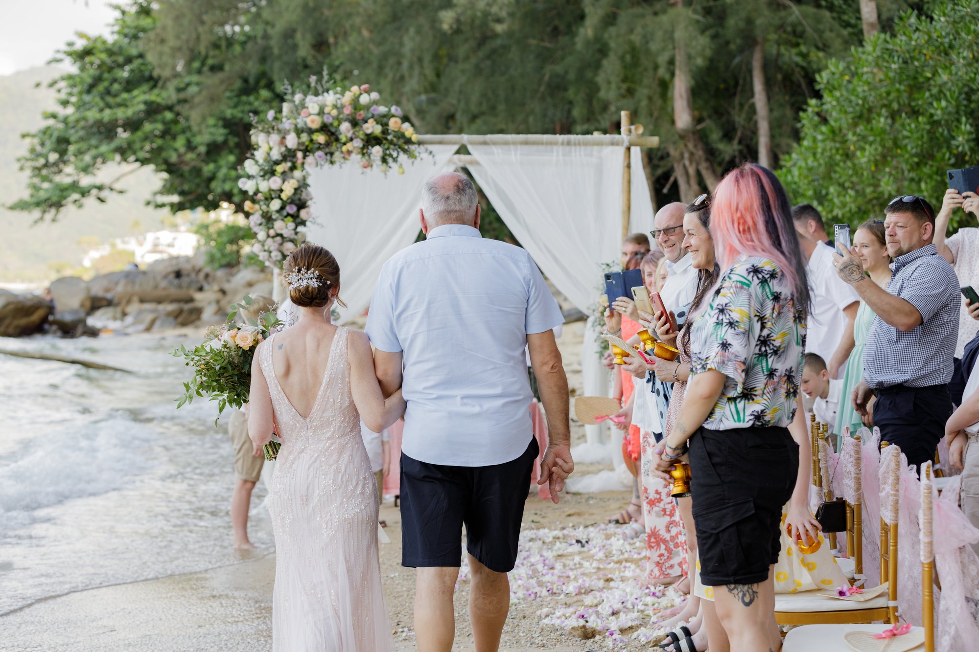 Amanda arriving down the petal-strewn aisle on her father John Jackson's arm at Hua Beach, guests standing on either side