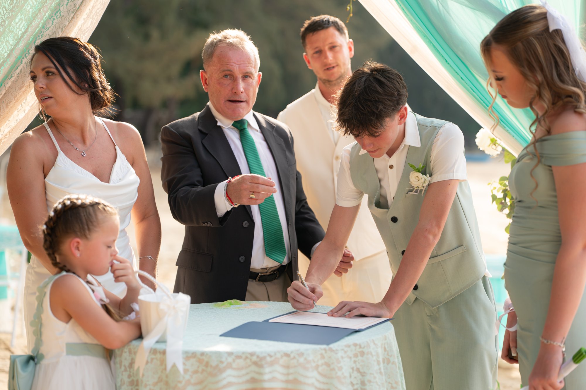 Nicky signing the marriage register at a small table on Hua Beach, Jade beside him and the celebrant looking on with the wedding party around them