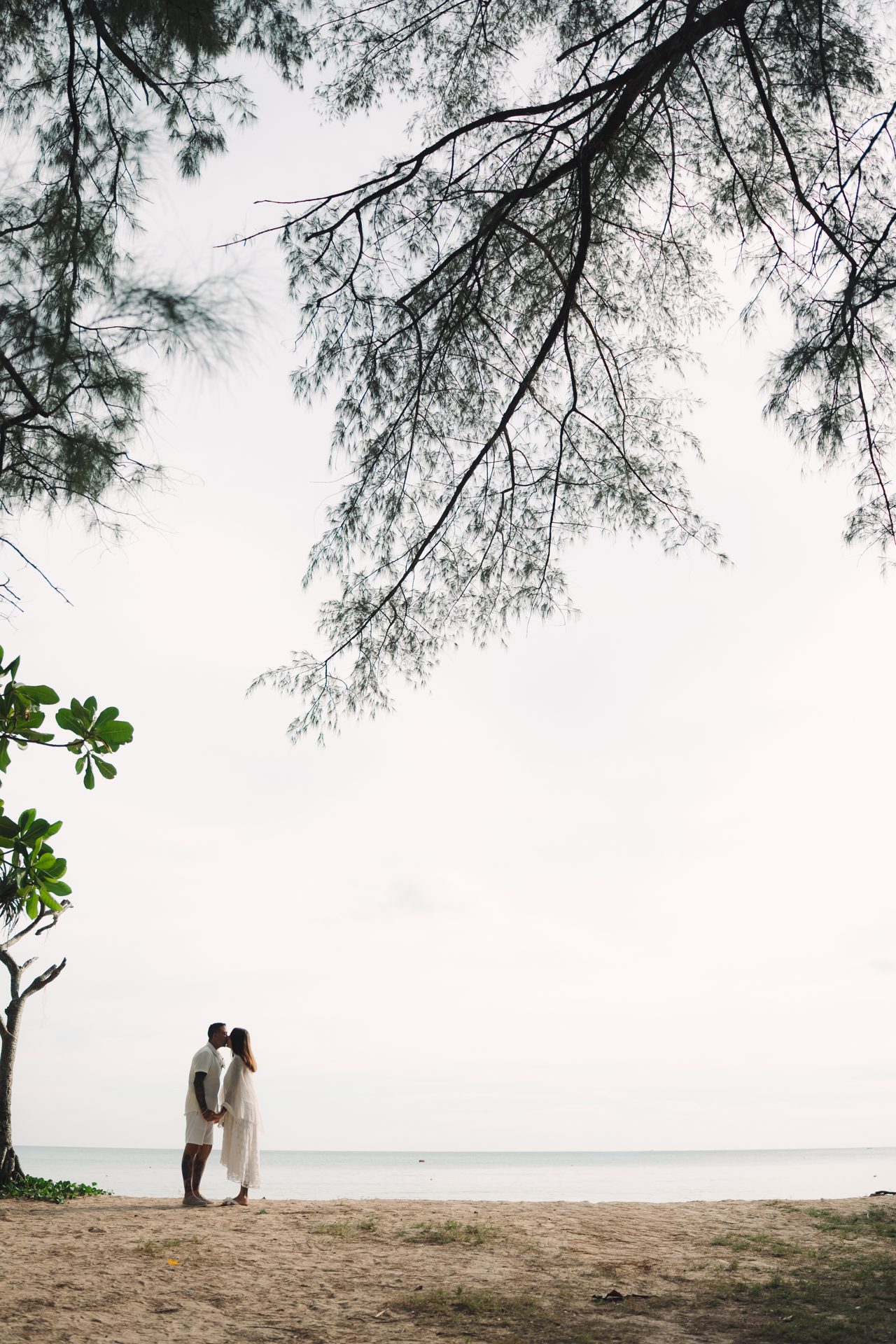 Distant silhouette of Joanne and Richard on the empty sand at Layan Beach, framed by overhanging trees