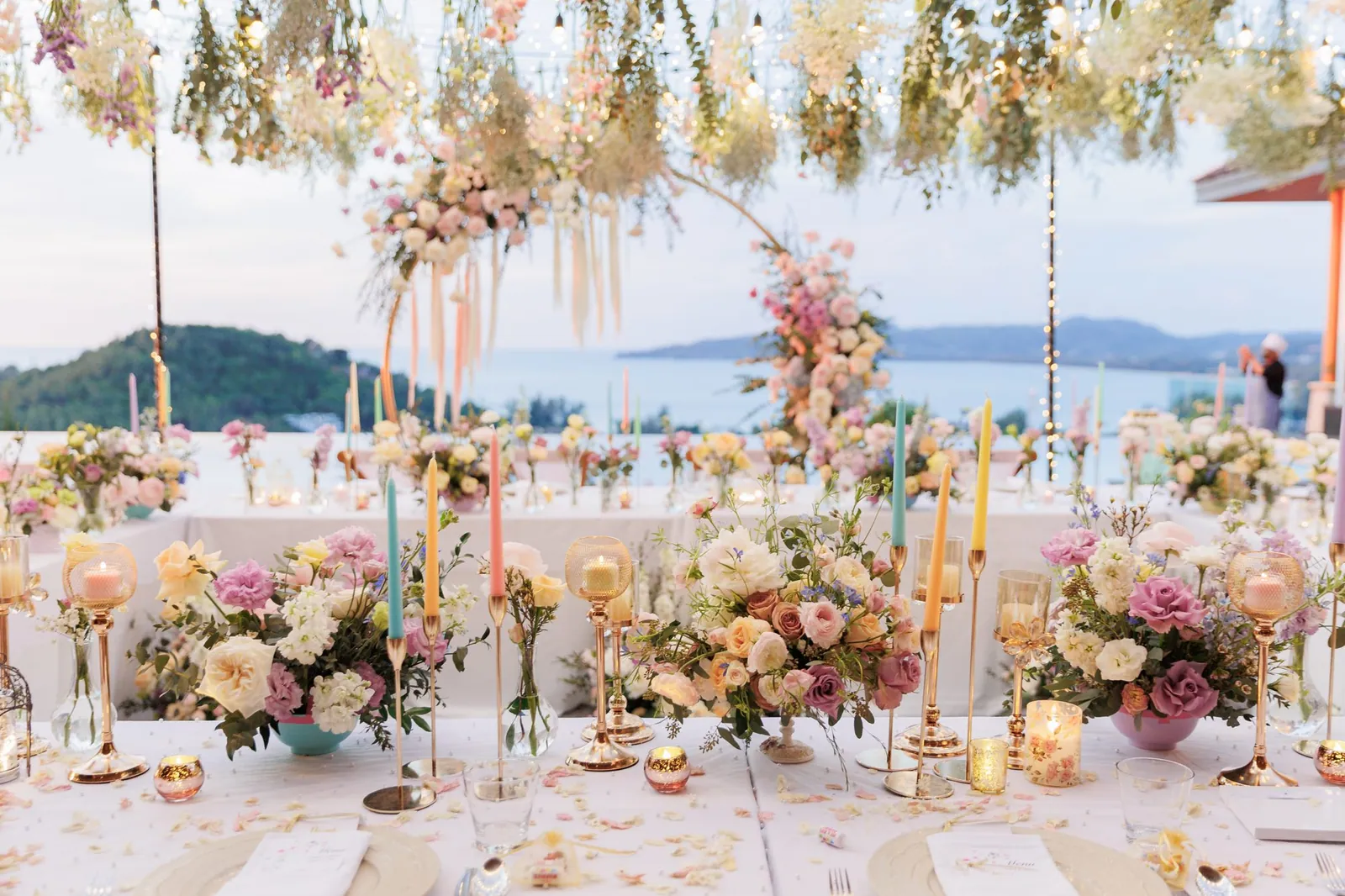 The long dinner table at Villa Baan Phu Prana set for twenty guests with pastel florals, tapered candles and the sea in the background at dusk
