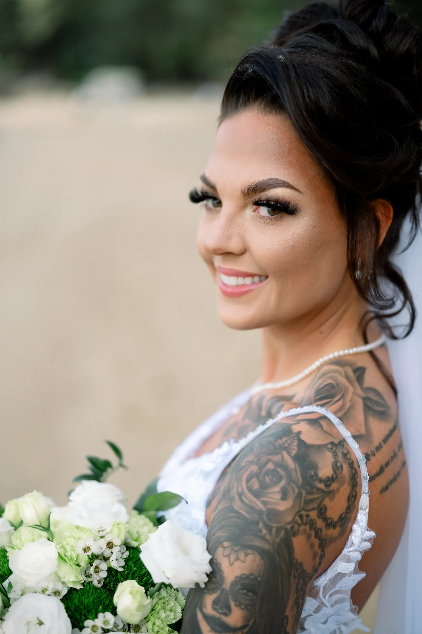 Rebecca in her lace wedding dress on Hua Beach, holding her white and green bouquet, tattoos visible along her arm, hair swept up