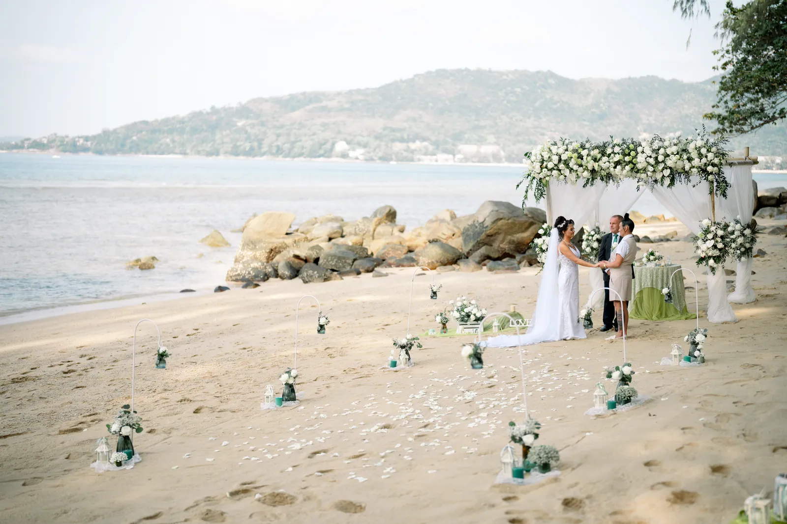 Wide view of the Hua Beach ceremony — Gemma and Rebecca standing beneath the bamboo arch, small lanterns lining a curved sand path, the sea and headland behind