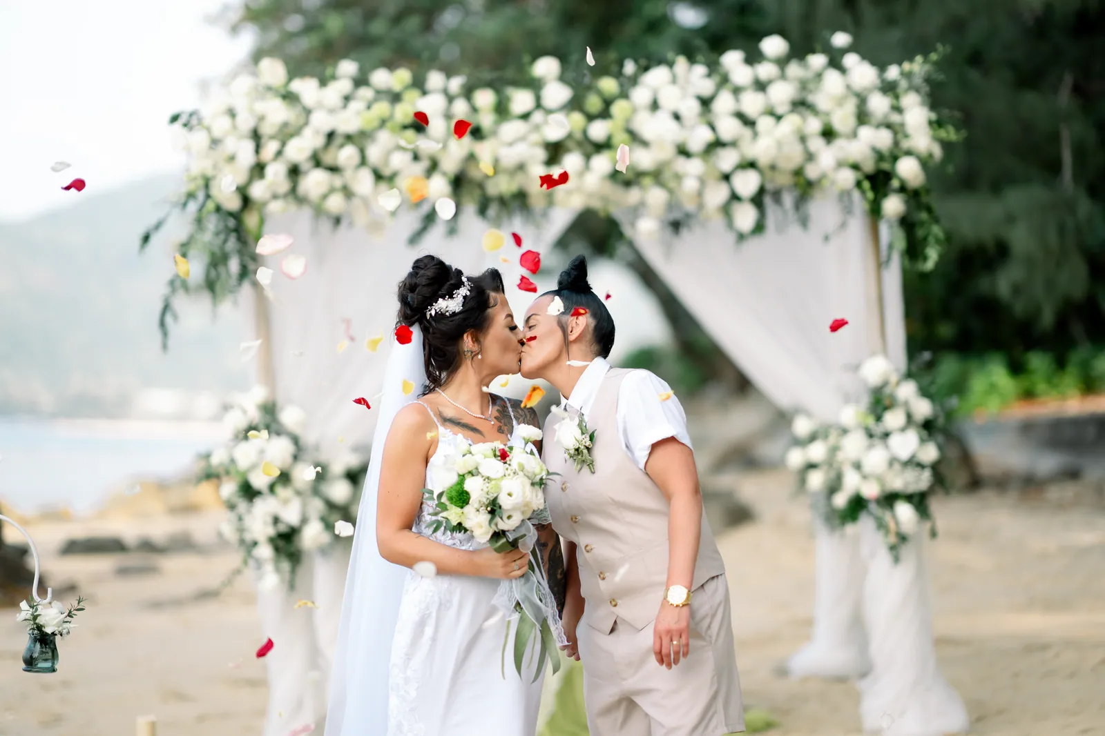 Gemma and Rebecca kissing under a lush bamboo flower arch on Hua Beach as red rose petals fall around them