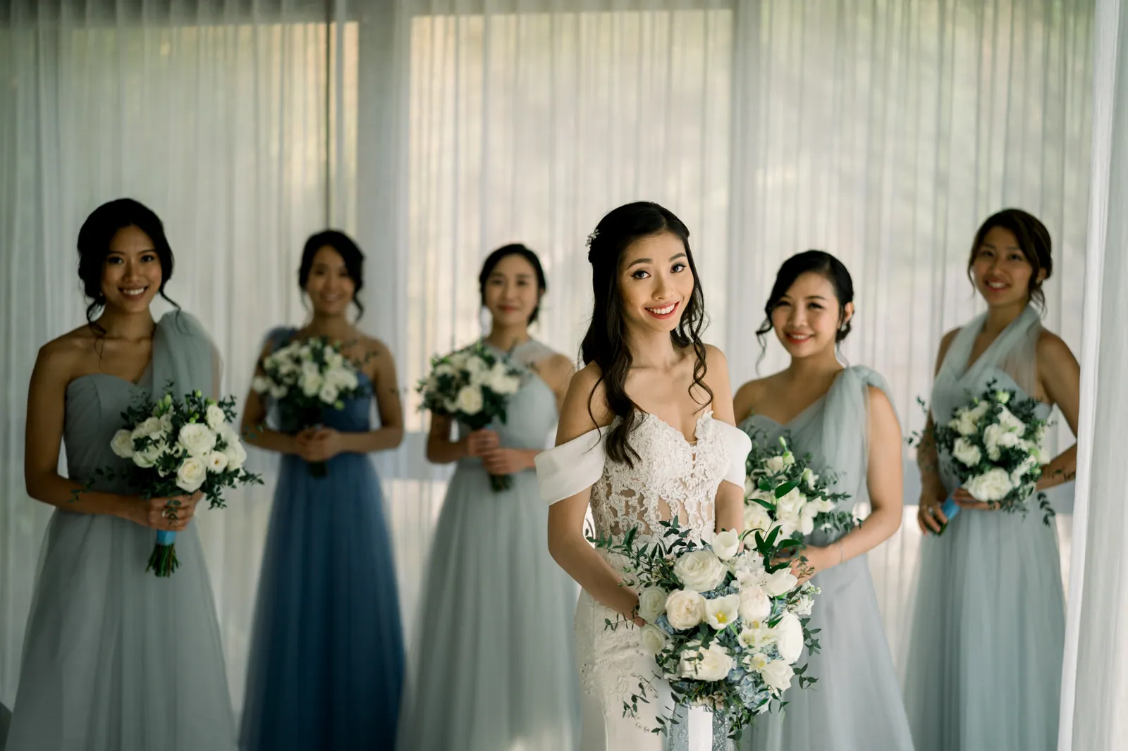 Eunice holding her white and dusty blue bouquet with her bridesmaids in matching dusty blue gowns