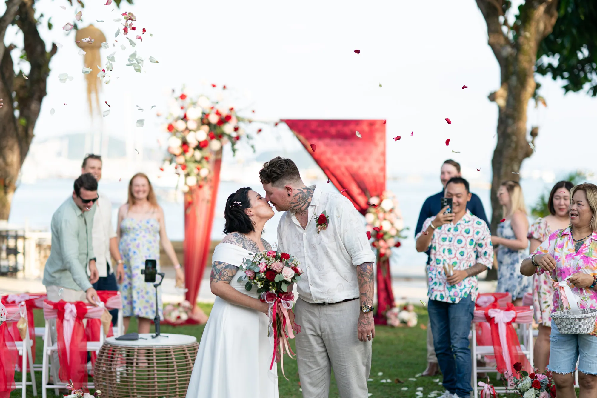 Louise & Daniel's elopement at Kan Eang @ Pier, Phuket — ceremony kiss beneath a red-and-pink floral arch with petals in the air.