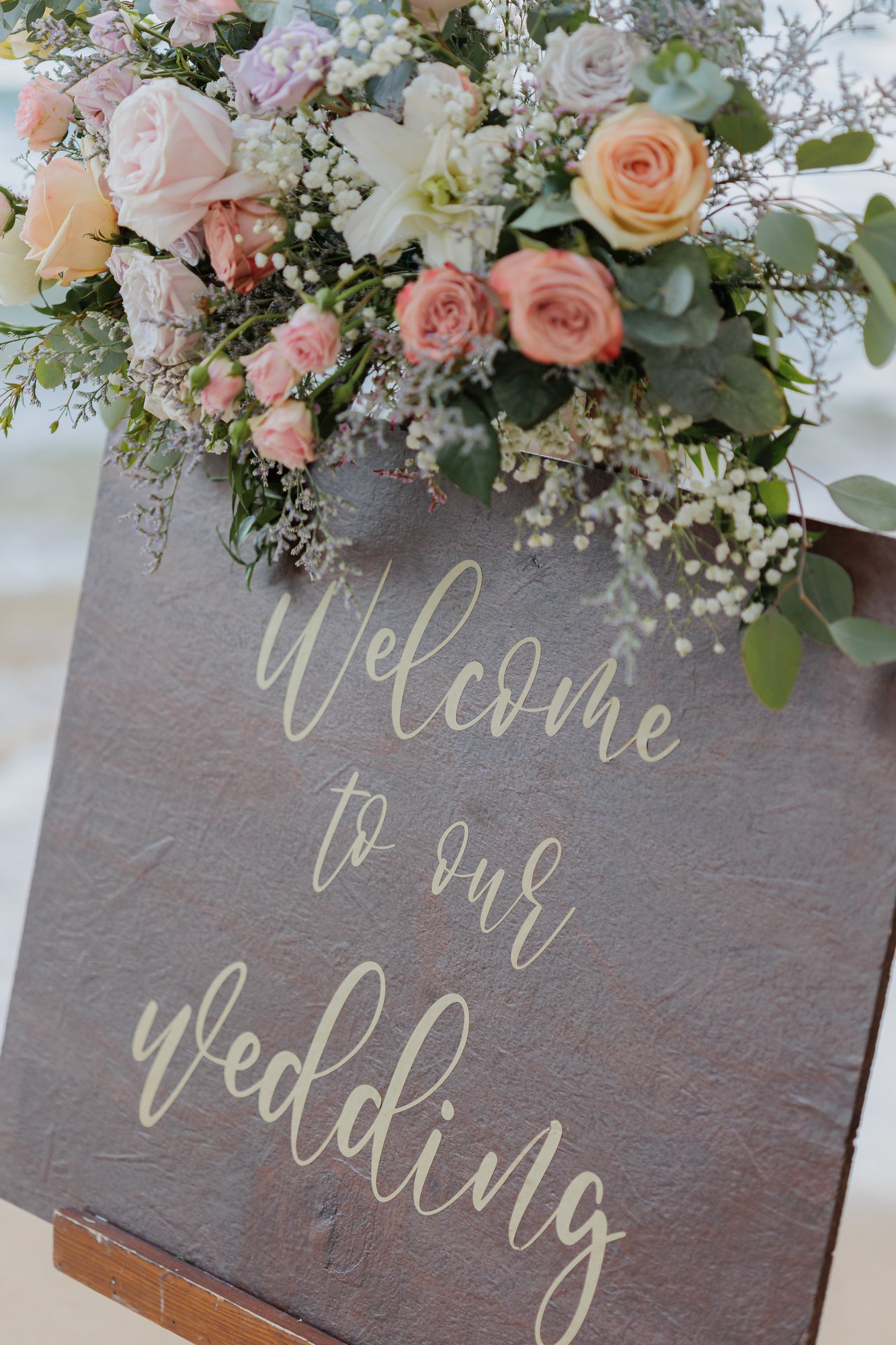 A hand-lettered 'Welcome to our wedding' sign in Amanda and John's blush-and-cream palette on the sand at Hua Beach