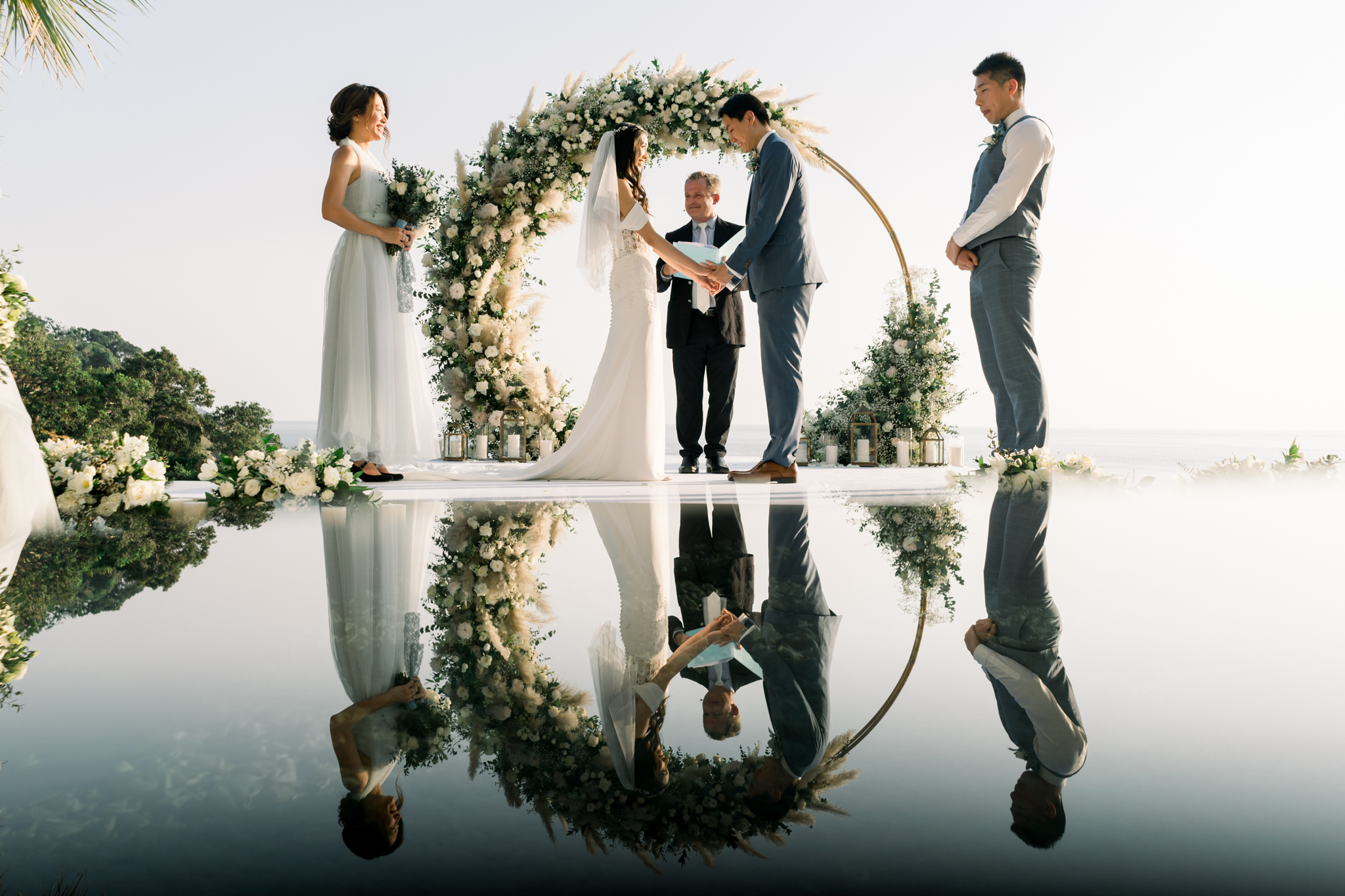 Eunice and Ryan holding hands under the circular gold floral arch on the platform over Villa Aquila's infinity pool, with Paul officiating