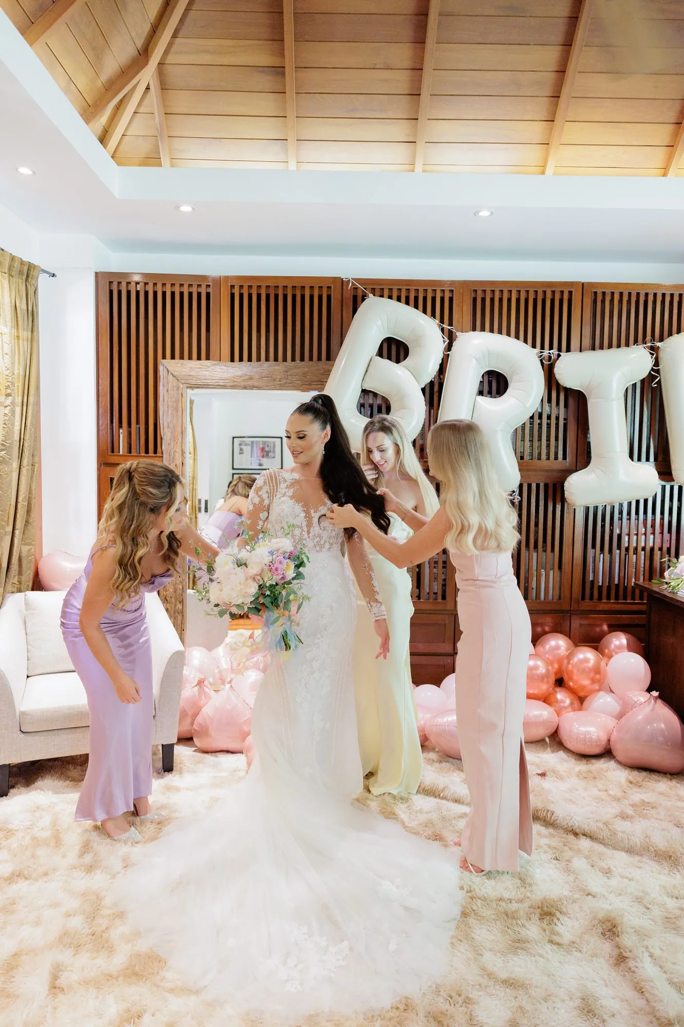Ashleigh in her wedding dress with her bridesmaids, standing beside BRIDE balloons in the villa's dressing area