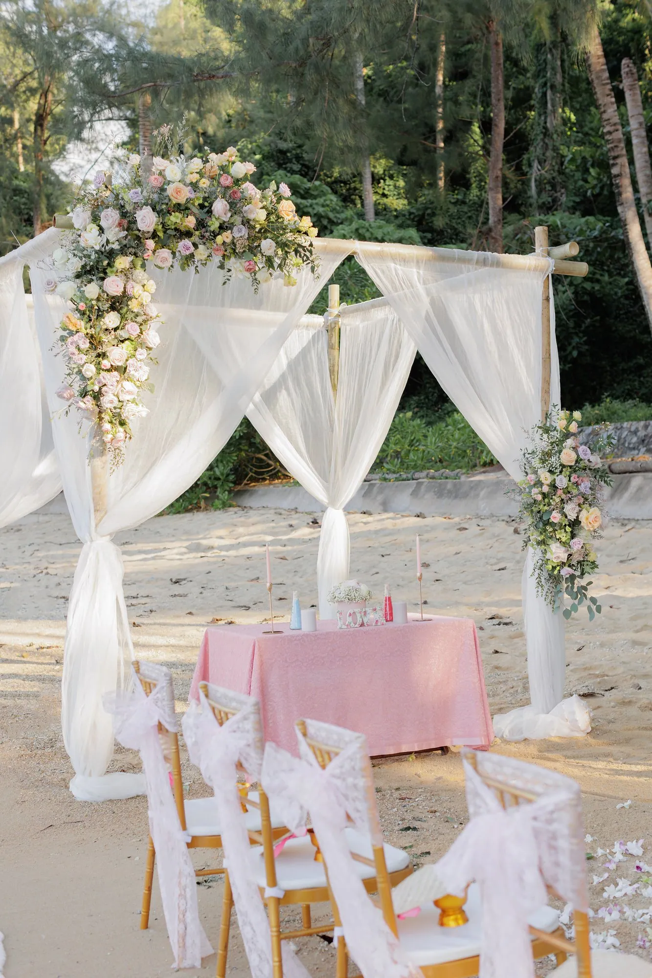 The bamboo ceremony arch dressed with white drapes, roses and eucalyptus at Hua Beach with pastel-styled aisle chairs