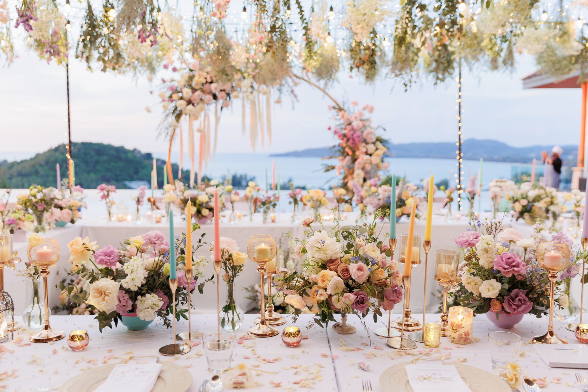 The long dinner table at Villa Baan Phu Prana set for twenty guests with pastel florals, tapered candles and the sea in the background at dusk