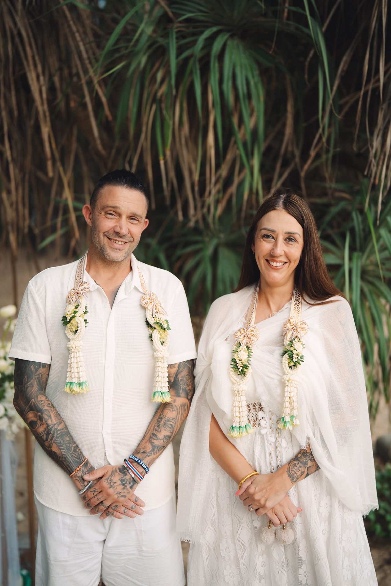 Joanne and Richard standing side by side in white after the ceremony, wearing floral garlands, framed by the trees at Layan Beach