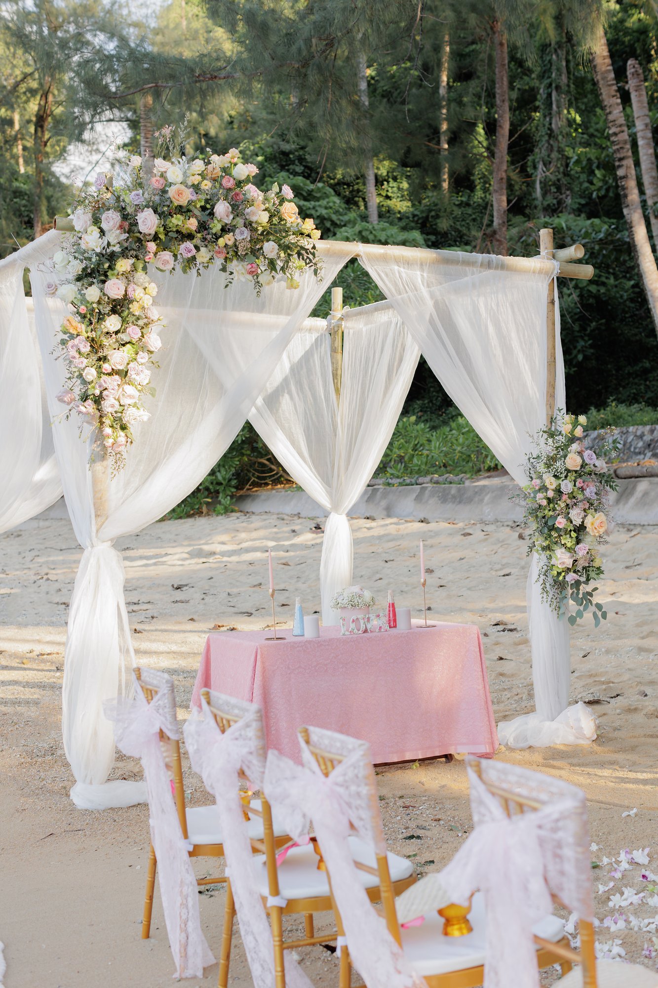 The bamboo ceremony arch dressed with white drapes, roses and eucalyptus at Hua Beach with pastel-styled aisle chairs