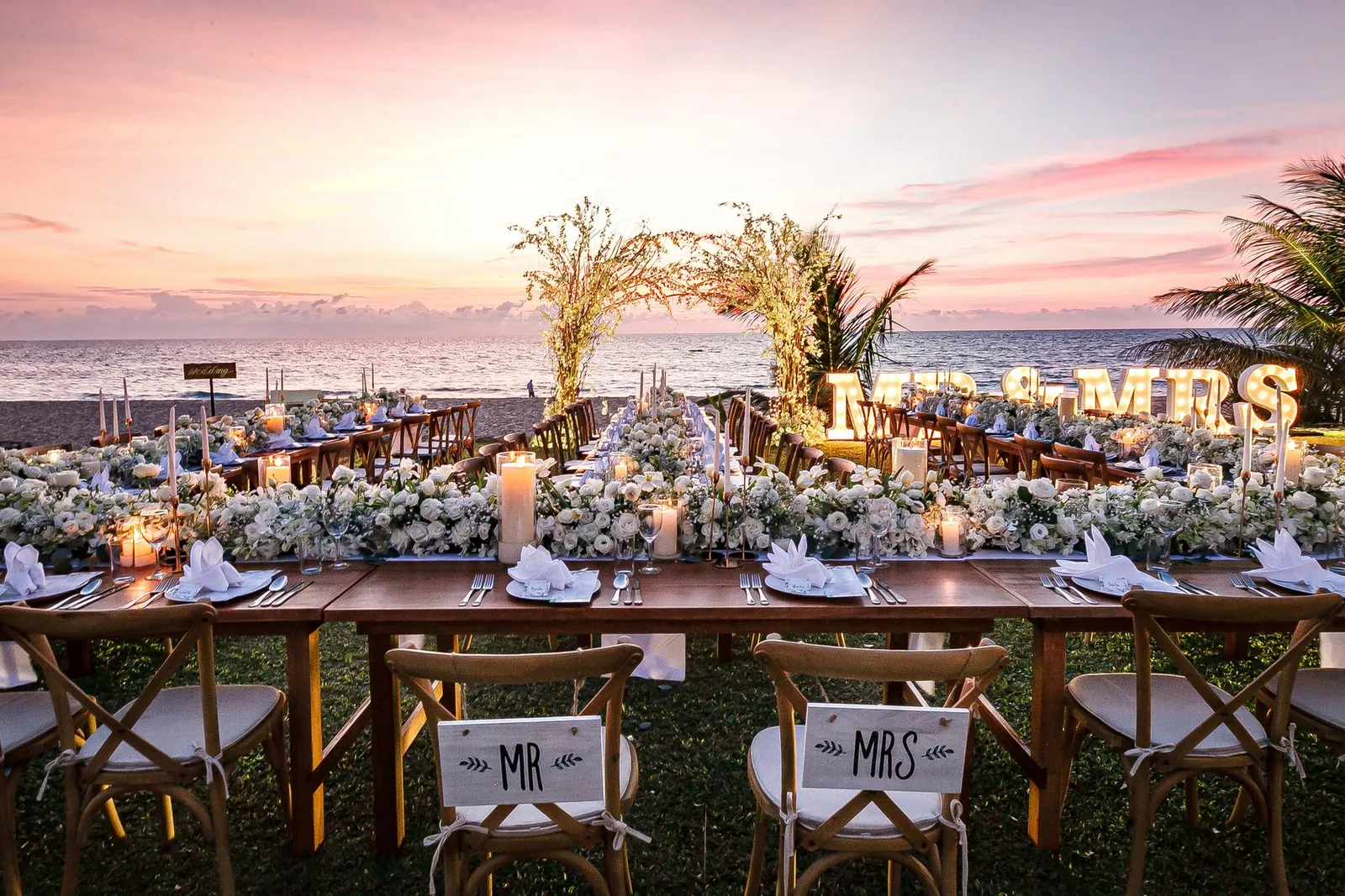 A long reception dinner table on the sand at sunset in Phuket, styled with white florals, candles and MR & MRS marquee letters, with a floral arch and the ocean behind