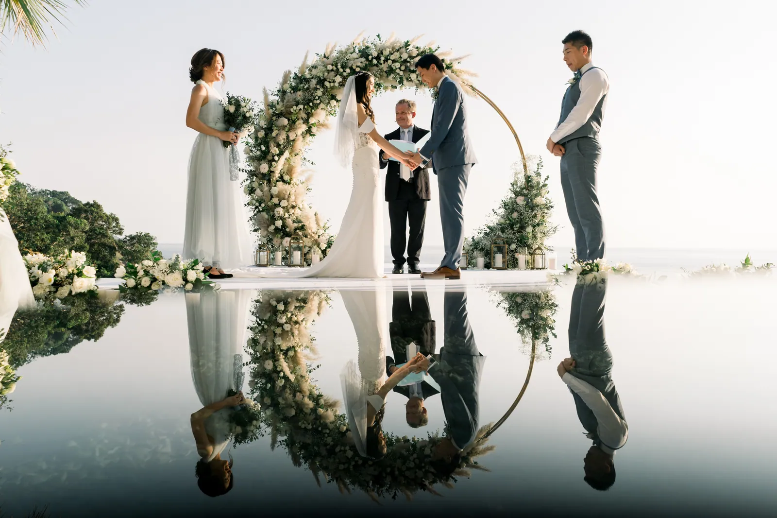 Eunice and Ryan holding hands under the circular gold floral arch on the platform over Villa Aquila's infinity pool, with Paul officiating