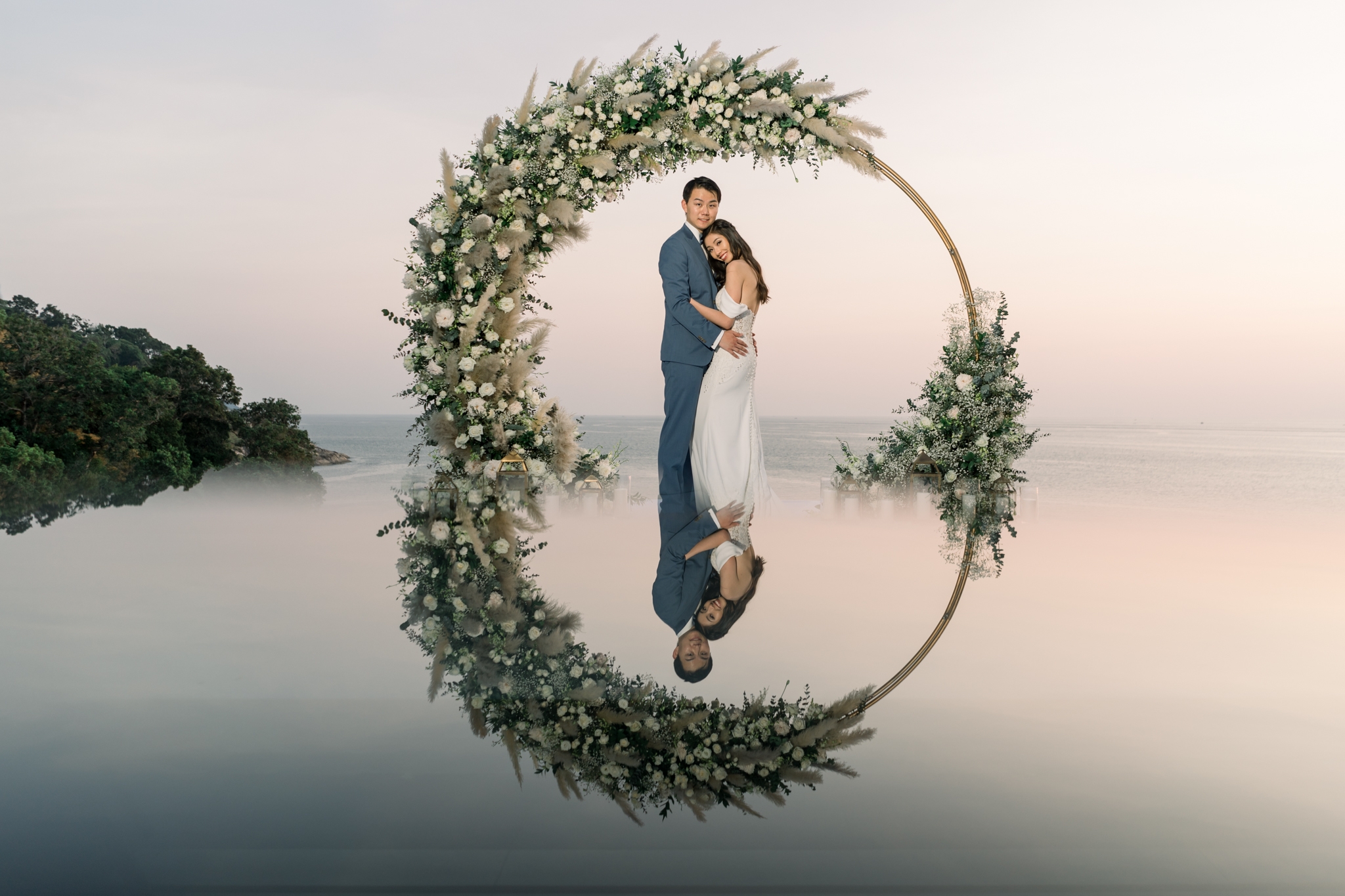 A bride and groom inside a circular floral arch, reflected on an infinity pool at dusk over the Andaman Sea — a Phuket wedding planned by Unique Phuket.