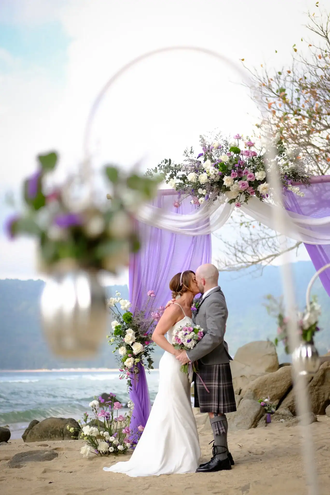 Bride and groom kissing under a purple floral ceremony arch during a beach elopement in Phuket